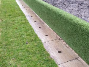 Artificial turf edging with wooden boundary featuring drilled holes for drainage or securing purposes.