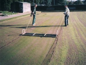 Two men scrubbing a pitch using a wide flat tool with a net in the middle