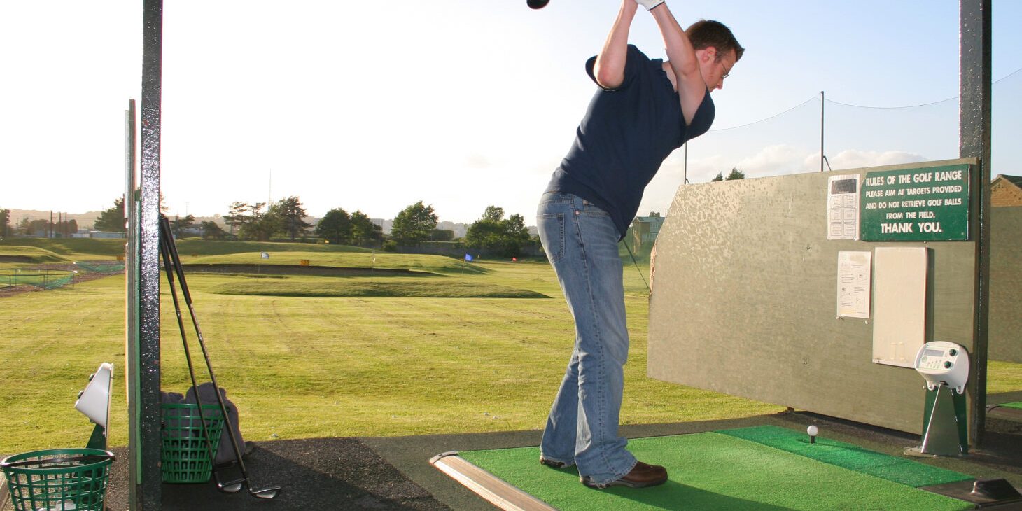Man with golfing pose on golf range Golfer practising a swing at a driving range, standing on a green mat with equipment and a scenic outdoor backdrop.