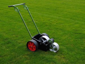 A lawn marking machine with red wheels, a black body, and metallic rollers, positioned on a vibrant green lawn.